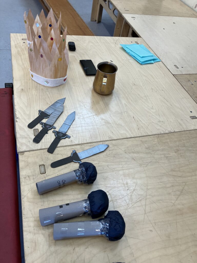 Three golden crowns, four silver and black cardboard daggers, four silver and black cardboard microphones, a golden tankard and two mobile phones on the stage at Great Leighs Primary. 