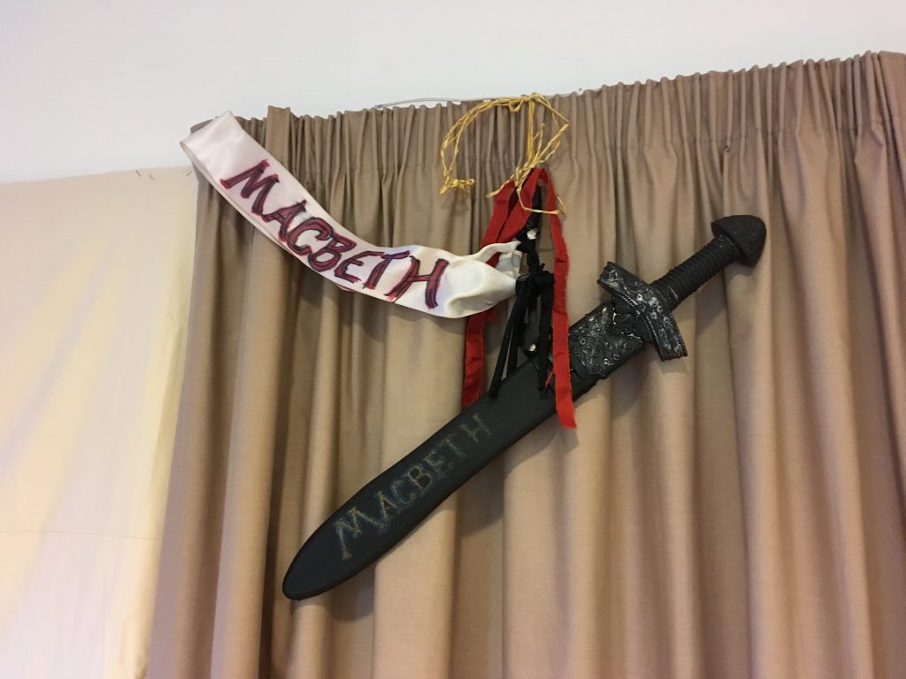 Black sword hanging by a red ribbon with a white wide ribbon attached with says Macbeth in black and red capital letters. It is set against a beige curtain.