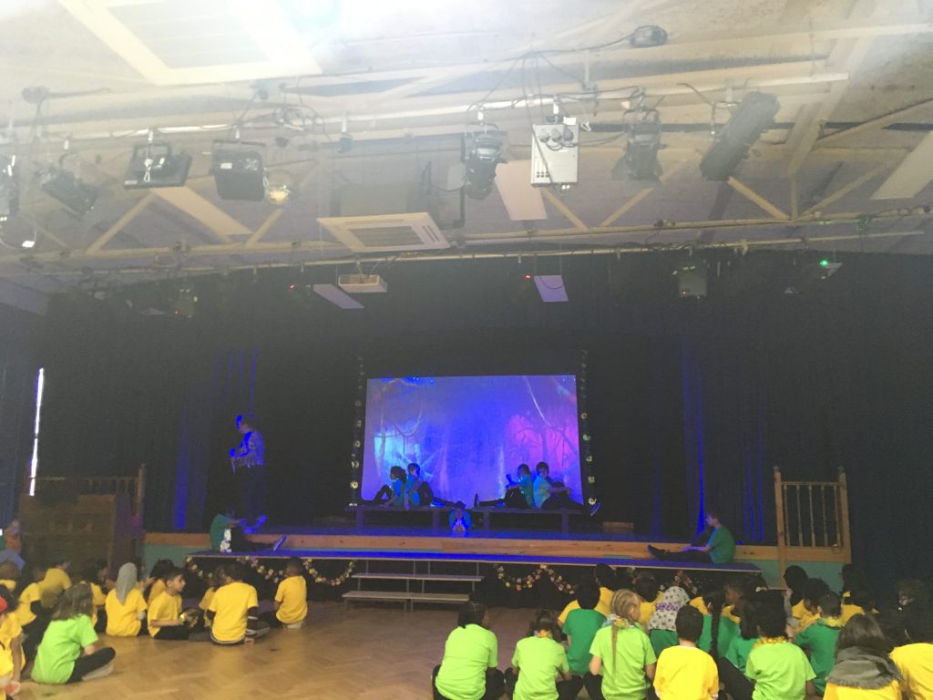 From the back of Anson Primary School hall, children sitting and facing the stage. They are wearing yellow and green T shirts. A scene on the stage from A Midsummer Night's Dream is taking place. It is a scene in the wood with a purple and red lighting effect. 