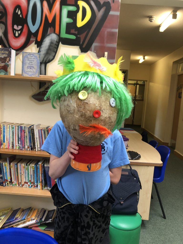 The prop severed head of Macbeth held by the child who made it in front of their own head. It is a grey ball with white button eyes, a red button nose, an orange feather mouth, green wool hair and a yellow crown. 