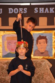 11 y old Hamlet about to kill 11 y old King Claudius who is wearing a red and gold crown. Portraits (drawn by children) on the wall behind.