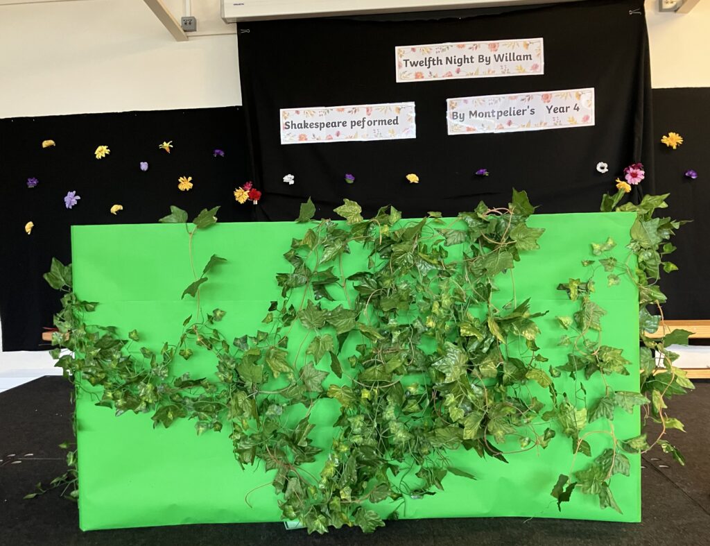 Green covered cardboard with leaves stapled to the front. Behind a black backdrop with flowers on it and the words: Twelfth Night by William Shakespeare, performed by Year 4.
