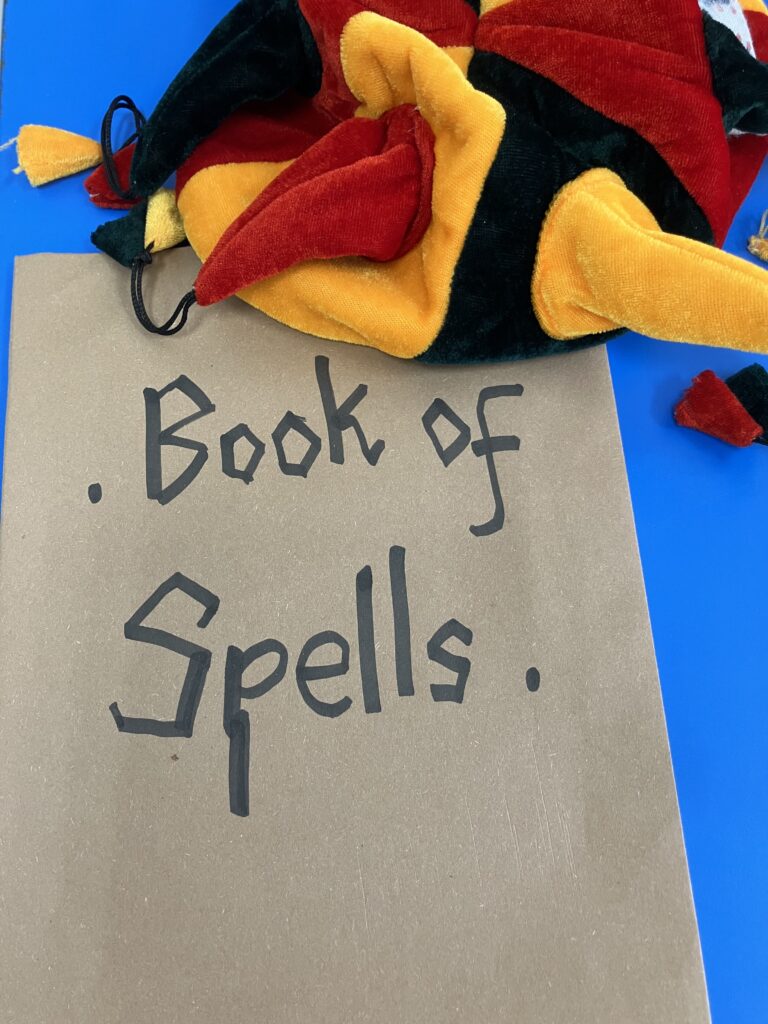 Jester hat (red, yellow and black) with a brown covered book that says Book of Spells in black ink.