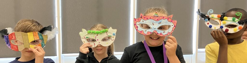 Head and shoulders of four children holding masks over their eyes. Decorated with paper and pen, the masks are orange, yellow, white and black. 