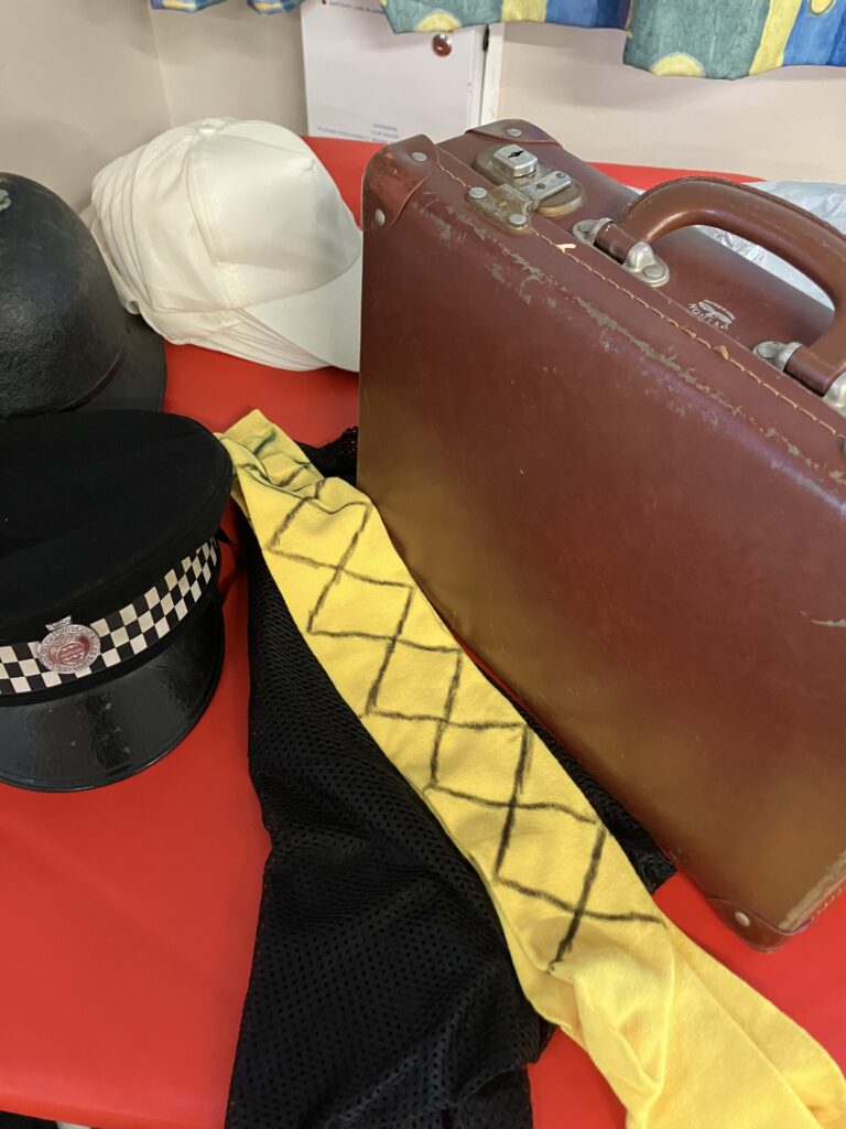 A small brown suitcase, a cross-gartered (in black) yellow sock, a black veil, two black police hats and a selection of white baseball caps on a red table.