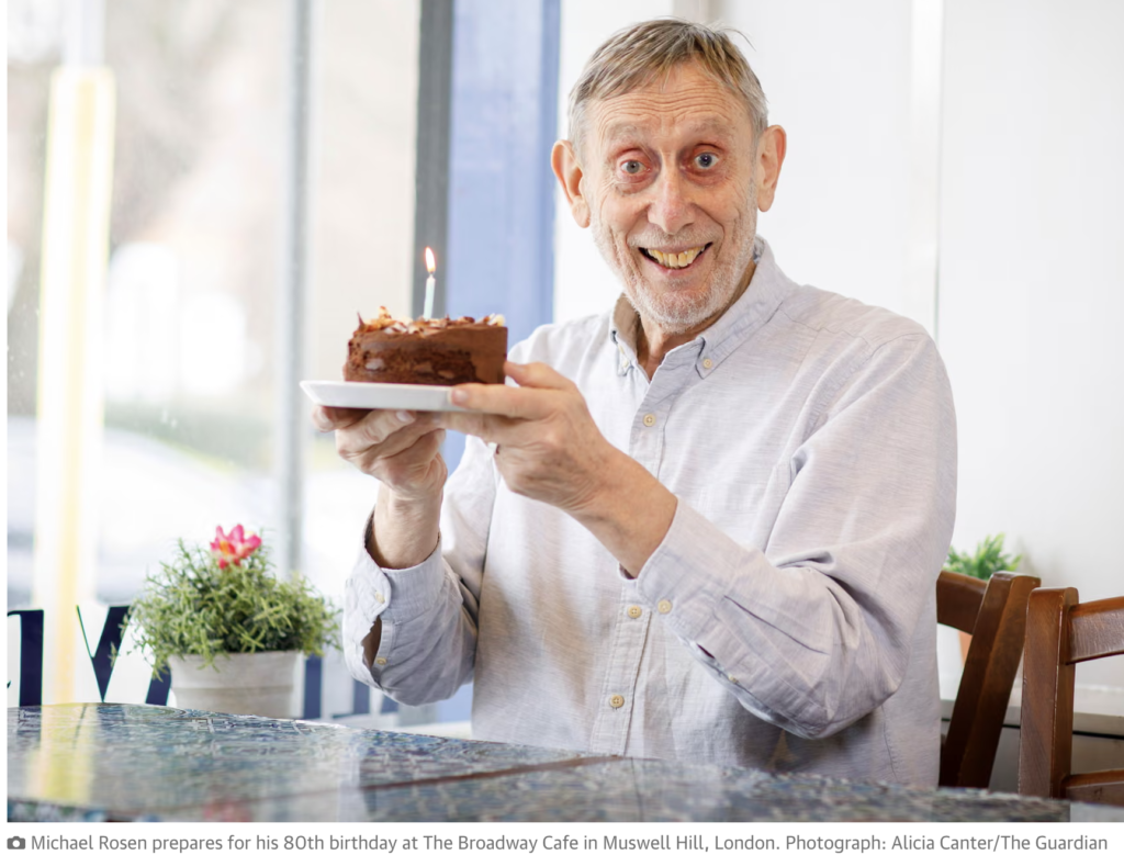 Photo courtesy of The Guardian of Michael Rosen in a white long sleeved shirt, smiling and holding up a small chocolate birthday cake with a candle on it.