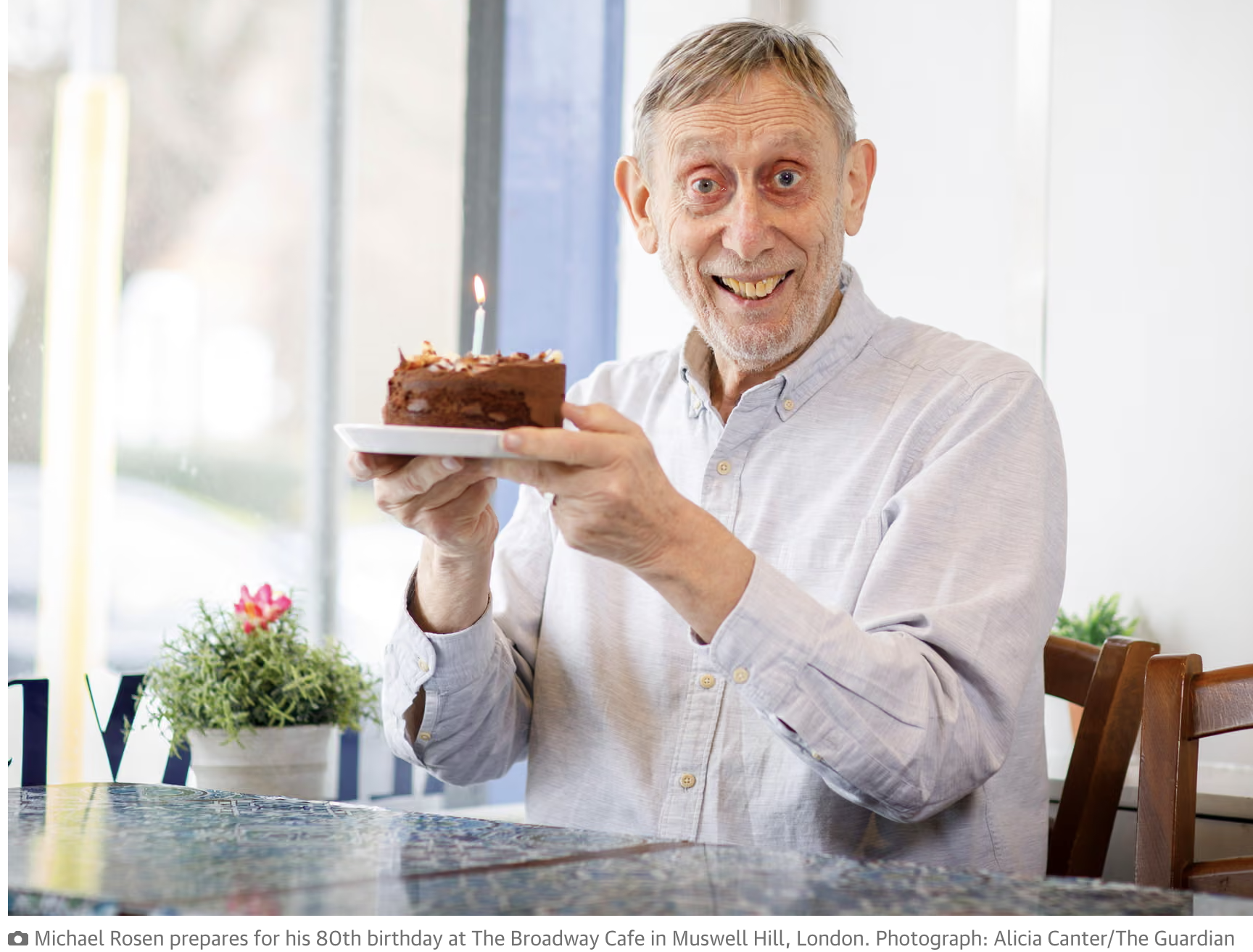 Photo courtesy of The Guardian of Michael Rosen in a white long sleeved shirt, smiling and holding up a small chocolate birthday cake with a candle on it.